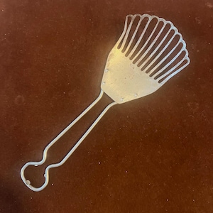 May include: A silver-coloured metal kitchen utensil with a fan-shaped head and a long handle. The head has multiple thin, parallel tines, and the handle has a looped end. The tool is likely vintage and made of metal.