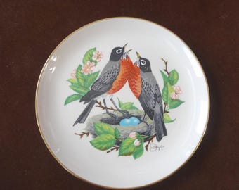 May include: A white porcelain plate with a gold rim features a painting of two robins perched on a nest with blue eggs. The robins are painted in shades of red, black, and gray, and the nest is surrounded by green leaves and pink flowers.