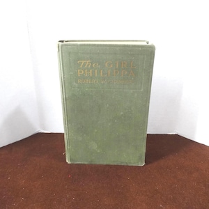 May include: A vintage hardcover book titled "The Girl Philippa" by Robert W. Chambers. The book has a faded green cover with gold lettering. The book is standing upright on a brown surface.