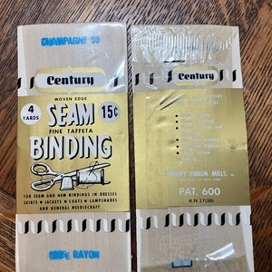 Könnte beinhalten: Zwei Packungen Century gewebter Saum-Bindeband aus feinem Taft. Die Packungen sind gold und weiß mit schwarzen und weißen Streifen. Der Text auf den Packungen lautet "Century Woven Edge Seam Binding Fine Taffeta 4 Yards 15c 100% Rayon Pat. 600 RN 17086".