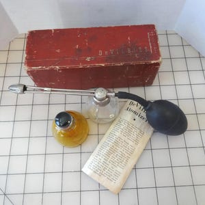 Puede incluir: Un juego de atomizador DeVilbiss antiguo con una caja roja, una botella de vidrio transparente y una botella de vidrio amarillo. El juego incluye una boquilla de pulverización de metal, una pera de goma negra y un papel impreso. La caja está etiquetada "DeVilbiss".