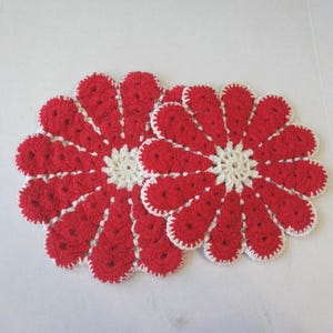 Op de afbeelding: Twee rood-witte gehaakte bloemvormige onderzetters. Elke onderzetter heeft een rood bloemblaadjesontwerp met een wit midden en witte rand. De onderzetters zijn handgemaakt en geschikt voor woondecoratie.