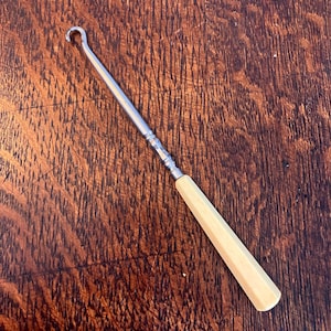 Puede incluir: Un ganchillo vintage con un eje de metal y un mango facetado de color crema. El gancho tiene un extremo curvado y está diseñado para tirar del hilo a través de bucles para crear tela. La herramienta se muestra sobre una superficie de madera.