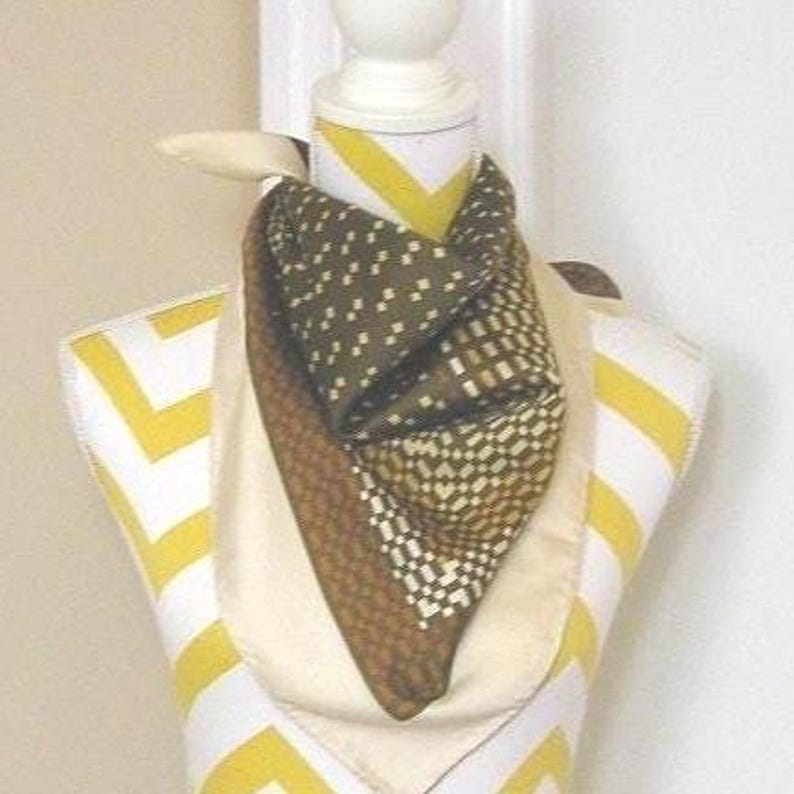 Peut inclure: Un foulard en soie avec des motifs g&eacute;om&eacute;triques dans les tons marron, vert olive et cr&egrave;me est drap&eacute; sur un mannequin. Le foulard est pli&eacute; pour cr&eacute;er un effet de superposition, mettant en valeur les diff&eacute;rents motifs et textures. Le mannequin a un design &agrave; rayures blanches et jaunes.