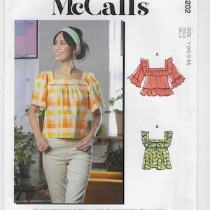 Könnte beinhalten: McCall's Schnittmuster M8202. Das Bild zeigt eine Frau in einem gelb-orange karierten Oberteil mit quadratischem Ausschnitt. Das Schnittmuster enthält mehrere Modelloptionen. Größe Y (XS-S-M).