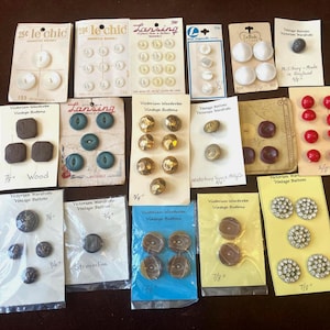 Puede incluir: Una colección de botones vintage en varias formas, tamaños y colores, exhibidos en sus embalajes originales. Los botones incluyen blanco, dorado, rojo y negro, algunos con detalles decorativos. La colección incluye botones de madera, metal y plástico.