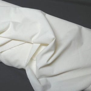 May include: A close-up shot of a folded, off-white fabric. The fabric has a smooth texture and appears to be a medium weight. The folds create shadows and highlights, adding depth to the image. The background is a dark gray.