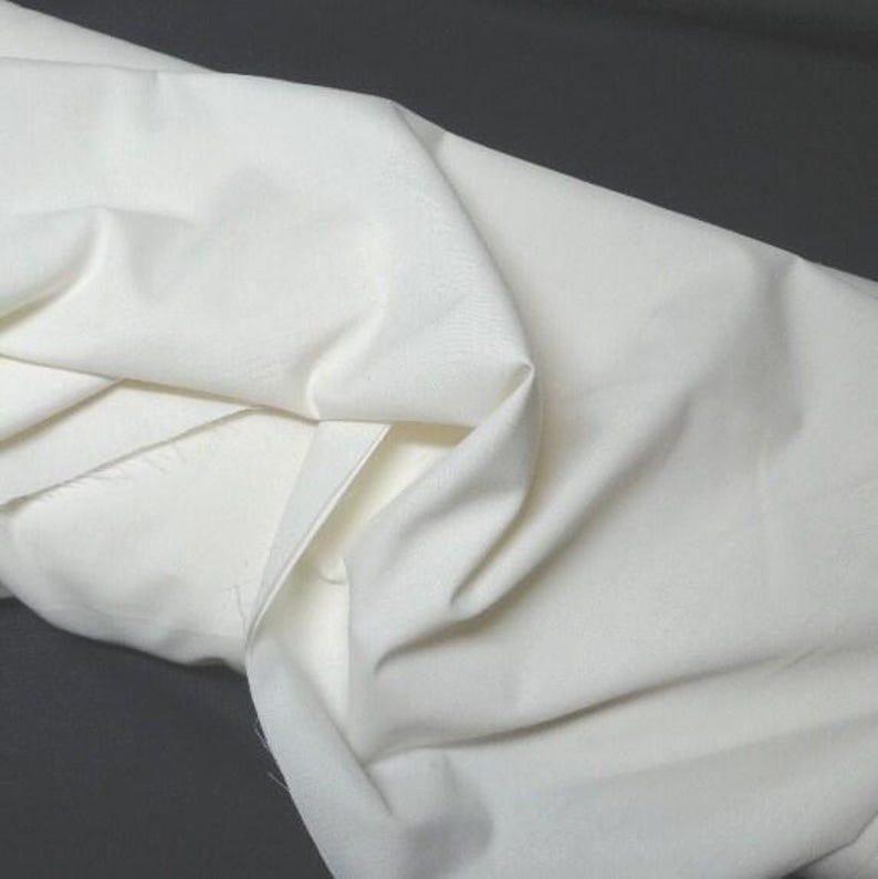 May include: A close-up shot of a folded, off-white fabric. The fabric has a smooth texture and appears to be a medium weight. The folds create shadows and highlights, adding depth to the image. The background is a dark gray.