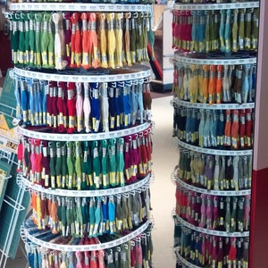 May include: A display of colorful embroidery floss in a craft store. The floss is arranged on circular, tiered racks, showcasing a wide array of hues, from vibrant reds and blues to softer greens and yellows. Each spool is labeled.