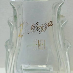 May include: A clear glass bottle with a textured surface. The bottle has a gold label with the text "Bellezza Lenel" on it.