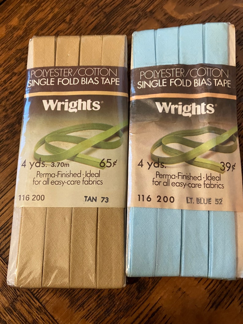 May include: Two packages of Wrights single fold bias tape. The package on the left is tan and the package on the right is light blue. Both packages are 4 yards long and are made of polyester and cotton. The tan package is 65 cents and the light blue package is 39 cents.