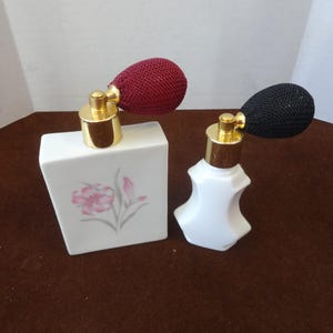 May include: Two vintage perfume atomizers. One is a white rectangular bottle with a pink floral design and a burgundy atomizer. The other is a white, shapely bottle with a black atomizer. Both have gold-colored metal accents.