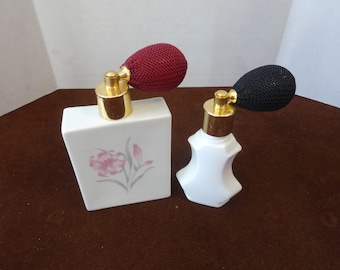 Puede incluir: Dos atomizadores de perfume vintage. Uno es un frasco rectangular blanco con un diseño floral rosa y un atomizador burdeos. El otro es un frasco blanco y con forma con un atomizador negro. Ambos tienen detalles metálicos dorados.