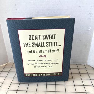 May include: A hardcover book titled "Don't Sweat the Small Stuff... and it's all small stuff." The book has a blue cover with a geometric pattern and a white rectangular panel with black text. The author's name, Richard Carlson, Ph.D., is at the bottom.