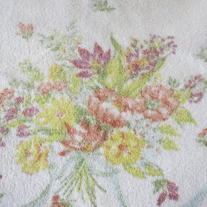 Könnte beinhalten: Weißes Handtuch mit floralem Muster. Das Design zeigt einen Blumenstrauß in Gelb-, Orange-, Rosa- und Grüntönen. Die Blumen sind dicht gepackt, mit einem weichen, strukturierten Aussehen. Das Handtuch hat eine klassische, Vintage-Ästhetik.
