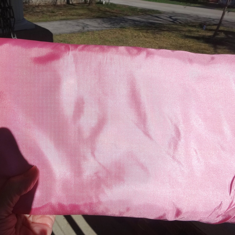 May include: A pink satin fabric, possibly a pillowcase or a piece of clothing, is held up in front of a natural background. The fabric is a solid, light pink color and appears to be smooth and shiny. The fabric is folded and has some wrinkles.