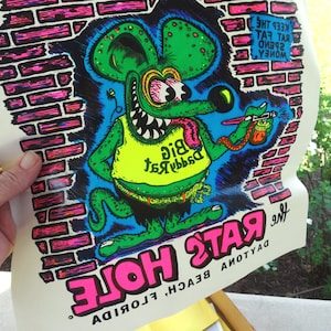 May include: A black and white illustration of a green rat with a yellow shirt that says "Daddy Rat" on it. The rat is holding a paint sprayer and a can of paint. The background is a brick wall. The text "The Rat's Hole" is written in pink letters at the bottom of the image. The text "Beach, Florida" is written in black letters below the pink text. The text "Keep the Rat Fat Spend Money" is written in black letters on the brick wall.