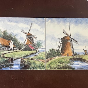 May include: Two ceramic tiles depicting Dutch windmills in a landscape. The tiles show windmills with brown blades and bodies, set against a backdrop of green fields, water, and a cloudy sky. The artwork evokes a sense of rural charm.
