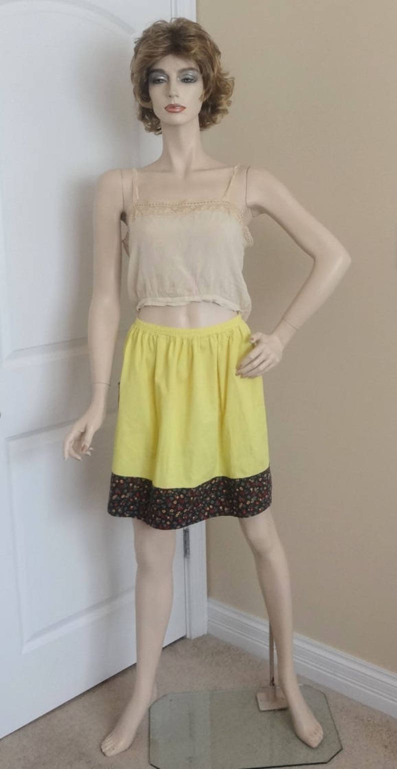 May include: A yellow skirt with a floral print hem. The skirt is worn by a mannequin with a light brown wig and a white lace top.