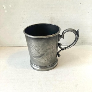 Könnte beinhalten: Ein Vintage-Becher aus silberfarbenem Metall mit dekorativem Griff und floraler Ätzung. Der Becher hat eine leicht konische Form und einen dunklen Innenraum. Der Griff ist verziert, und das florale Design verleiht dem Stück einen Hauch von Eleganz.