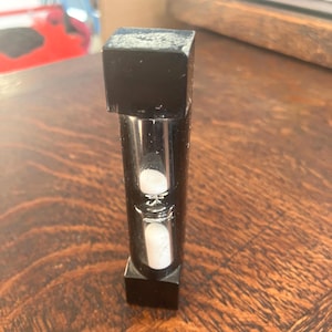May include: A black hourglass with a square top and bottom. The clear glass tube contains white sand. The hourglass is a decorative item, likely used for timing or as a unique desk accessory.