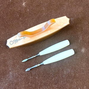 May include: An antique manicure set featuring a rectangular, light-colored case with a curved handle and decorative floral accents. Two ivory-colored cuticle pushers with metal tips are also present, suggesting a vintage beauty tool collection.