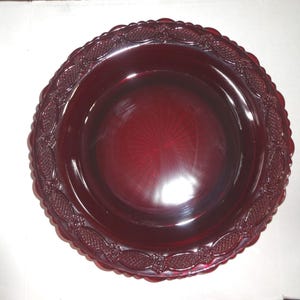 May include: A deep red glass plate with a decorative, embossed border. The plate has a circular shape with a slightly raised center and a diameter of approximately 25 cm. The border features a repeating pattern of stylized floral and geometric elements.
