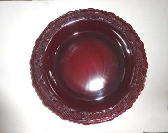 May include: A deep red glass plate with a decorative, embossed border. The plate has a circular shape with a slightly raised center and a diameter of approximately 10 inches. The border features a repeating pattern of stylized floral and geometric elements.