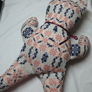 May include: A plush teddy bear made from a pink, white, and blue patterned fabric. The bear has a burgundy ribbon tied around its neck.