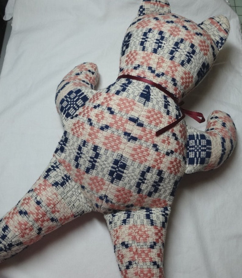 May include: A plush teddy bear made from a pink, white, and blue patterned fabric. The bear has a burgundy ribbon tied around its neck.