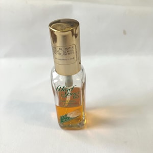 May include: A small, clear glass perfume bottle with a gold-colored cap. The bottle contains a yellow liquid and has the words "Wind Song" printed on the front. The label also includes the words "NATURAL SPRAY" and "VALUE".