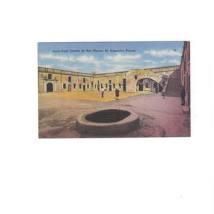 May include: Vintage postcard featuring the Court Yard of Castillo de San Marcos in St. Augustine, Florida. The image shows a large courtyard with a circular structure in the foreground and a historic building in the background. The sky is blue with some clouds.