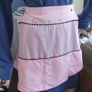 May include: A pink and white striped apron with black trim and a scalloped edge. The apron has a single pocket and a button closure.