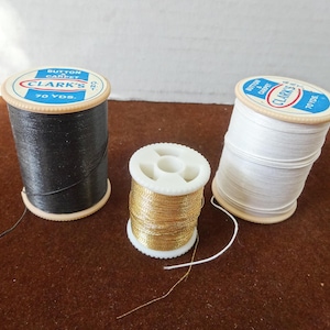 Könnte beinhalten: Drei Garnrollen: schwarz, gold und weiß. Die schwarzen und weißen Spulen sind mit "CLARK'S BUTTON CARPET 70 YDS." beschriftet. Das goldene Garn befindet sich auf einer kleineren, weißen Spule. Die Fäden liegen auf einer braunen Oberfläche.