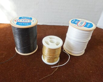 Puede incluir: Tres carretes de hilo: negro, dorado y blanco. Los carretes negro y blanco están etiquetados como "CLARK'S BUTTON CARPET 70 YDS." El hilo dorado está en un carrete blanco más pequeño. Los hilos están sobre una superficie marrón.