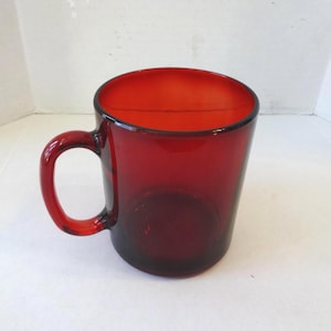 May include: A ruby red glass mug with a curved handle. The mug has a cylindrical shape with a slightly flared top. The glass is transparent, allowing light to pass through, and the handle is securely attached.