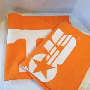 Pode incluir: Dois itens de tecido laranja com gráficos brancos. Um exibe um grande "T" branco, o outro apresenta um design branco com uma estrela. Os itens estão dobrados, sugerindo que são toalhas ou artigos de tecido semelhantes.