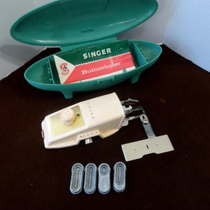 Op de afbeelding: Een vintage Singer knoopsgatmachine met een wit kunststof behuizing en een groene kunststof koffer. De koffer heeft een label met de tekst "Singer Knoopsgatmachine".