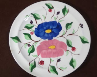 May include: A white ceramic plate with a floral design. The plate features two flowers, one blue and one pink, with yellow centers. The flowers are surrounded by green leaves and stems.