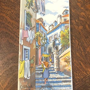 May include: Rectangular ceramic tile featuring a colorful illustration of a street scene in Lisbon, Portugal. The artwork depicts a woman carrying a yellow cloth on her head, walking up steps. The tile has the words "LISBOA ANTIGA" printed on it.
