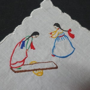 Könnte beinhalten: Weißes, besticktes Taschentuch mit zwei Figuren in traditioneller Kleidung. Die Figuren sind mit rotem, blauem, gelbem und grünem Faden gestickt. Das Taschentuch hat einen gewellten Rand.