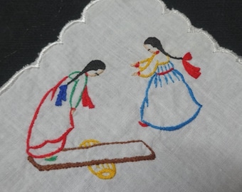 May include: White embroidered handkerchief featuring two figures in traditional attire. The figures are depicted in red, blue, yellow, and green thread. The handkerchief has a scalloped edge.