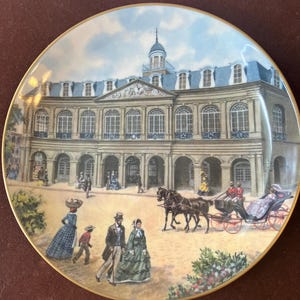 May include: A round decorative porcelain plate with a gold-trimmed edge featuring a detailed illustration of a large classical building with arched windows and a cupola. In the foreground, people in period clothing walk near a horse-drawn carriage.