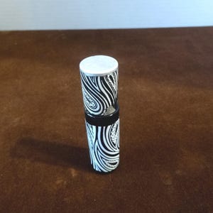 May include: A cylindrical perfume bottle with a black and white zebra stripe pattern. The bottle has a silver cap and a black band near the middle. The design features curved lines and paisley-like details.