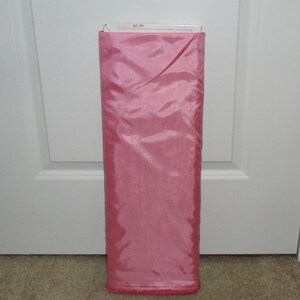 May include: A roll of pink fabric, likely for sewing or crafting. The fabric is a solid, light pink color with a shiny, smooth texture. The top of the roll has a white label with text. The fabric is neatly folded and ready for use.