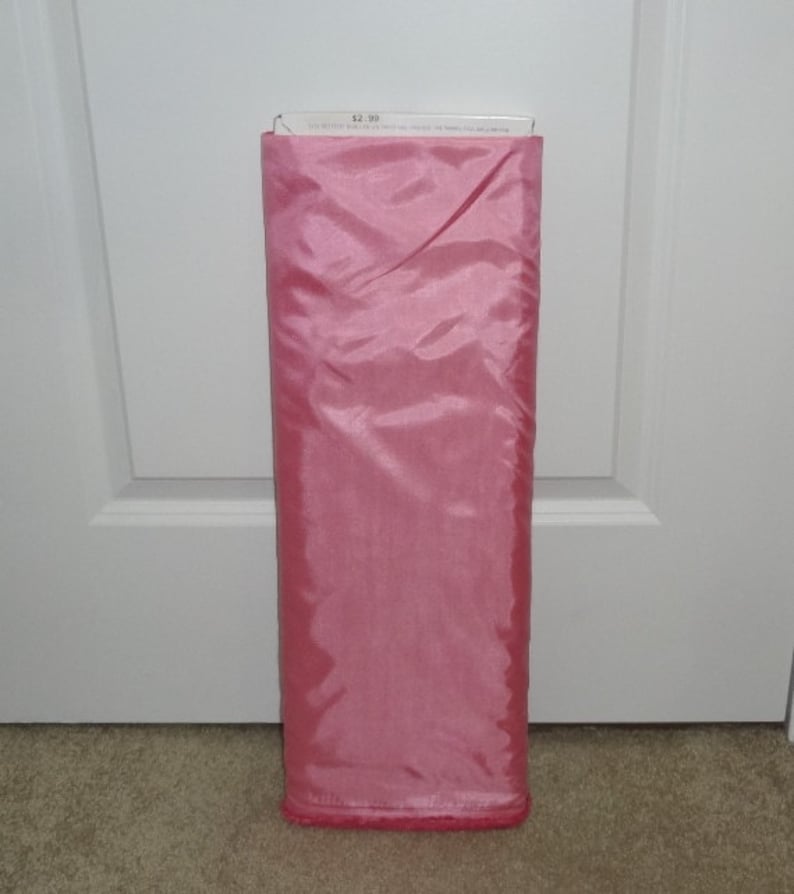 May include: A roll of pink fabric, likely for sewing or crafting. The fabric is a solid, light pink color with a shiny, smooth texture. The top of the roll has a white label with text. The fabric is neatly folded and ready for use.