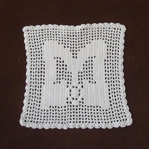 以下が含まれることがあります： 中央に大きな透かし彫りの文字「M」が施された、正方形の白いかぎ針編みのドイリー。縁はスカラップで、ドイリーは茶色の背景に配置されています。この手作りのアイテムは、家の装飾に適しています。