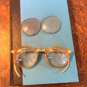 May include: A pair of round, translucent amber-colored eyeglasses with clear lenses and two separate, circular, dark-tinted replacement lenses positioned above the frame. The eyeglasses feature a classic bridge and curved temple tips.