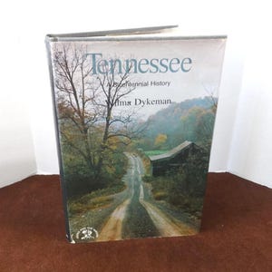 May include: A book titled "Tennessee: A Bicentennial History" by Wilma Dykeman. The cover features a photograph of a winding dirt road through a forest with a small wooden building in the distance.