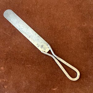 May include: A vintage metal spatula with a rectangular blade and a looped handle. The handle is made of wire and has text etched into it. The spatula has a worn, aged appearance, suggesting it has been used.